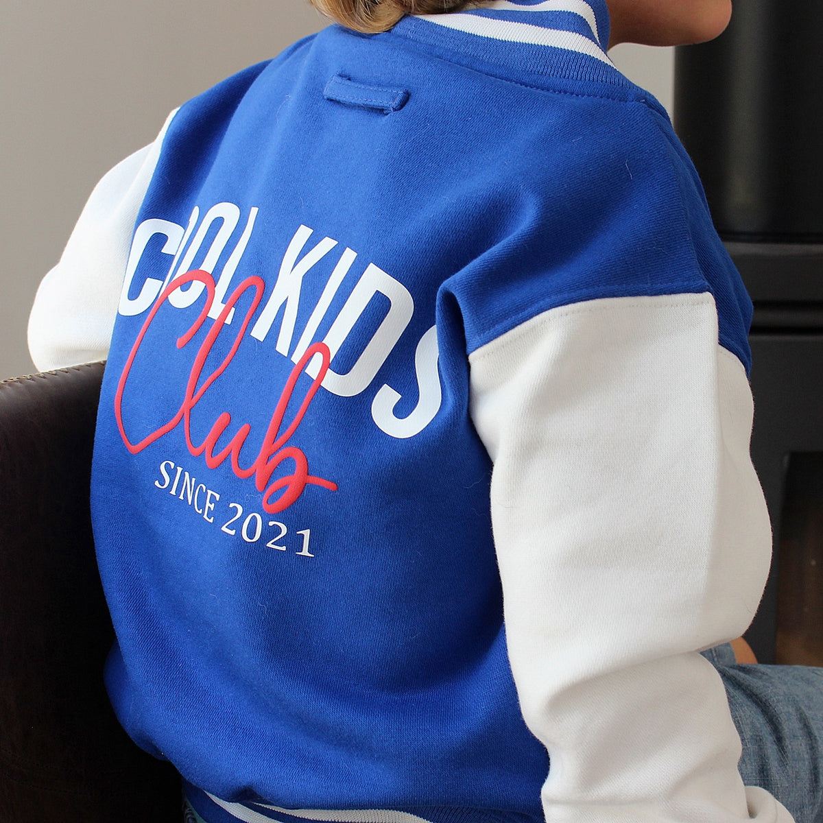 A child wearing a royal blue and white varsity-style jacket, viewed from the back. The jacket features 'COOL KIDS Club SINCE 2021' printed on the back in white and red lettering.