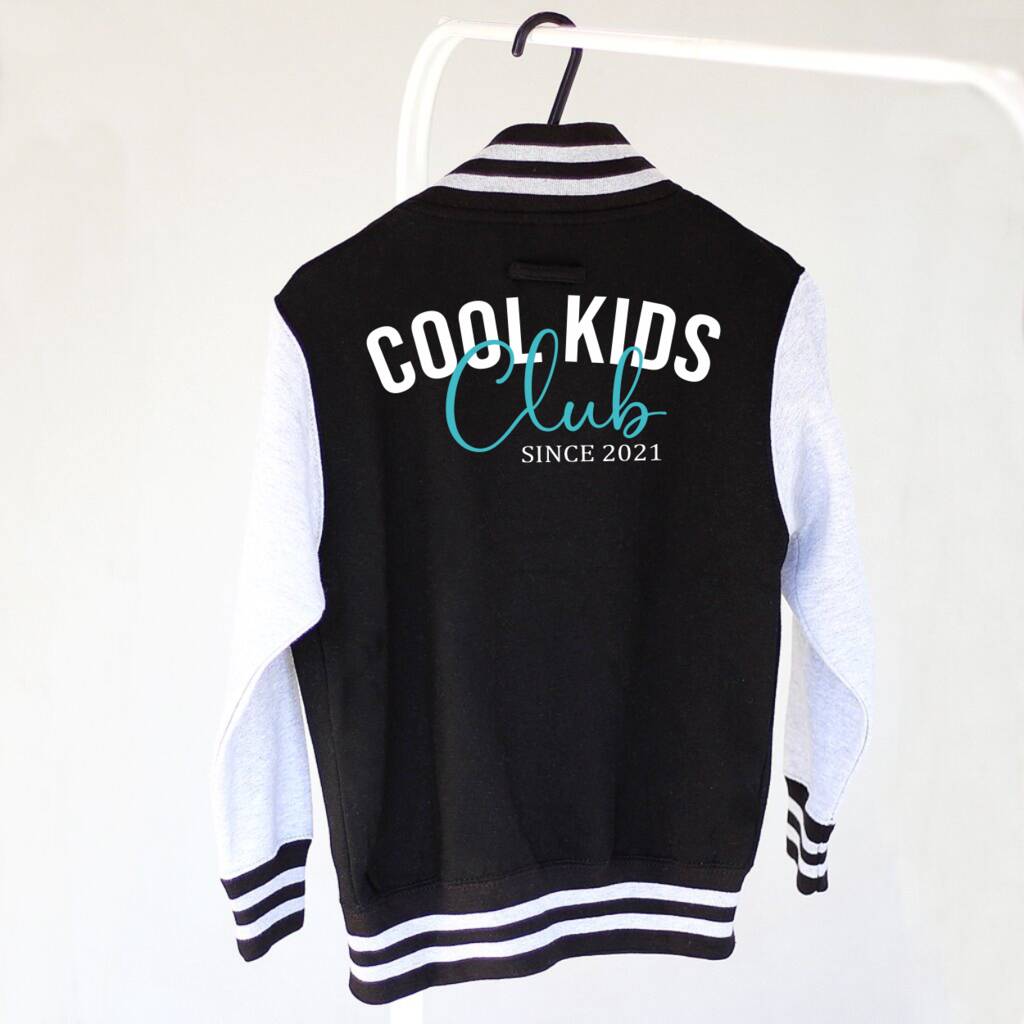 A black and white varsity jacket hanging on a white hanger against a white wall. The back of the jacket features 'COOL KIDS Club SINCE 2021' printed in white and teal text.