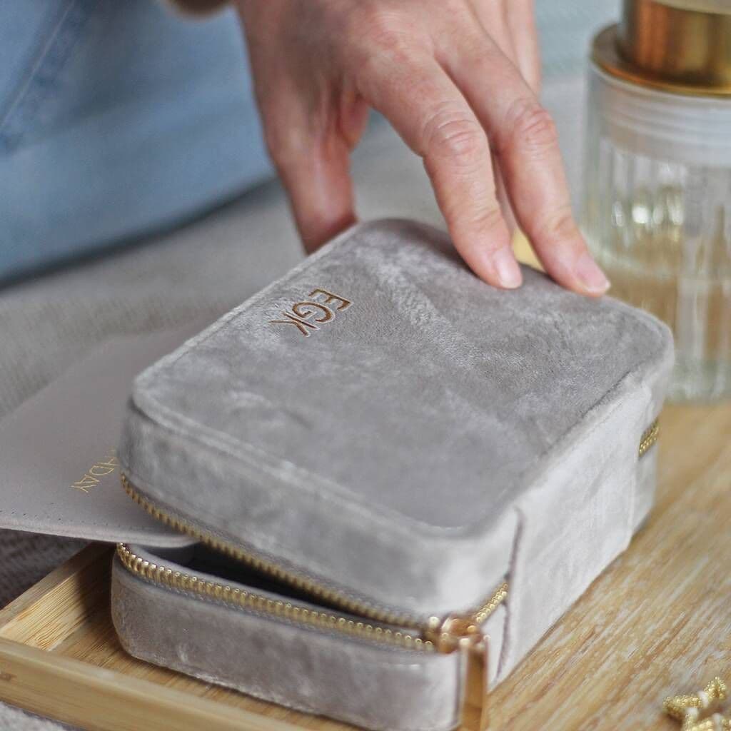 Gray velvet jewelry box with gold zipper on a wooden surface, hand reaching towards it.