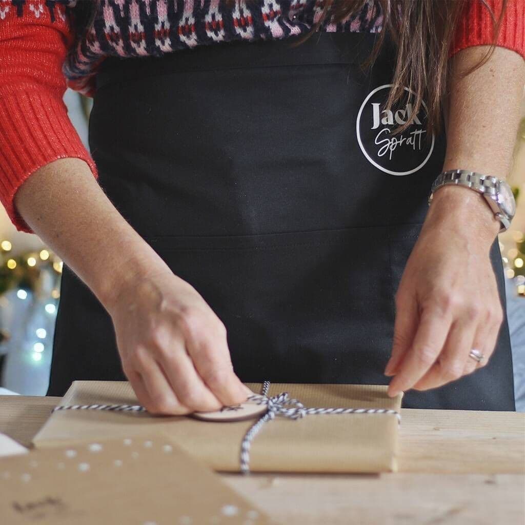 Person in a black apron with 'Jack Spratt' logo wrapping a gift on a wooden table.