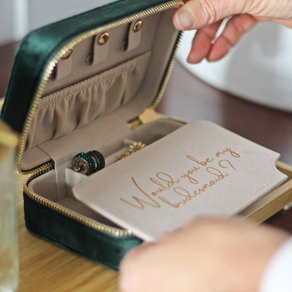 Jewelry box with a card inside asking 'Would you be my bridesmaid?' on a wooden surface.