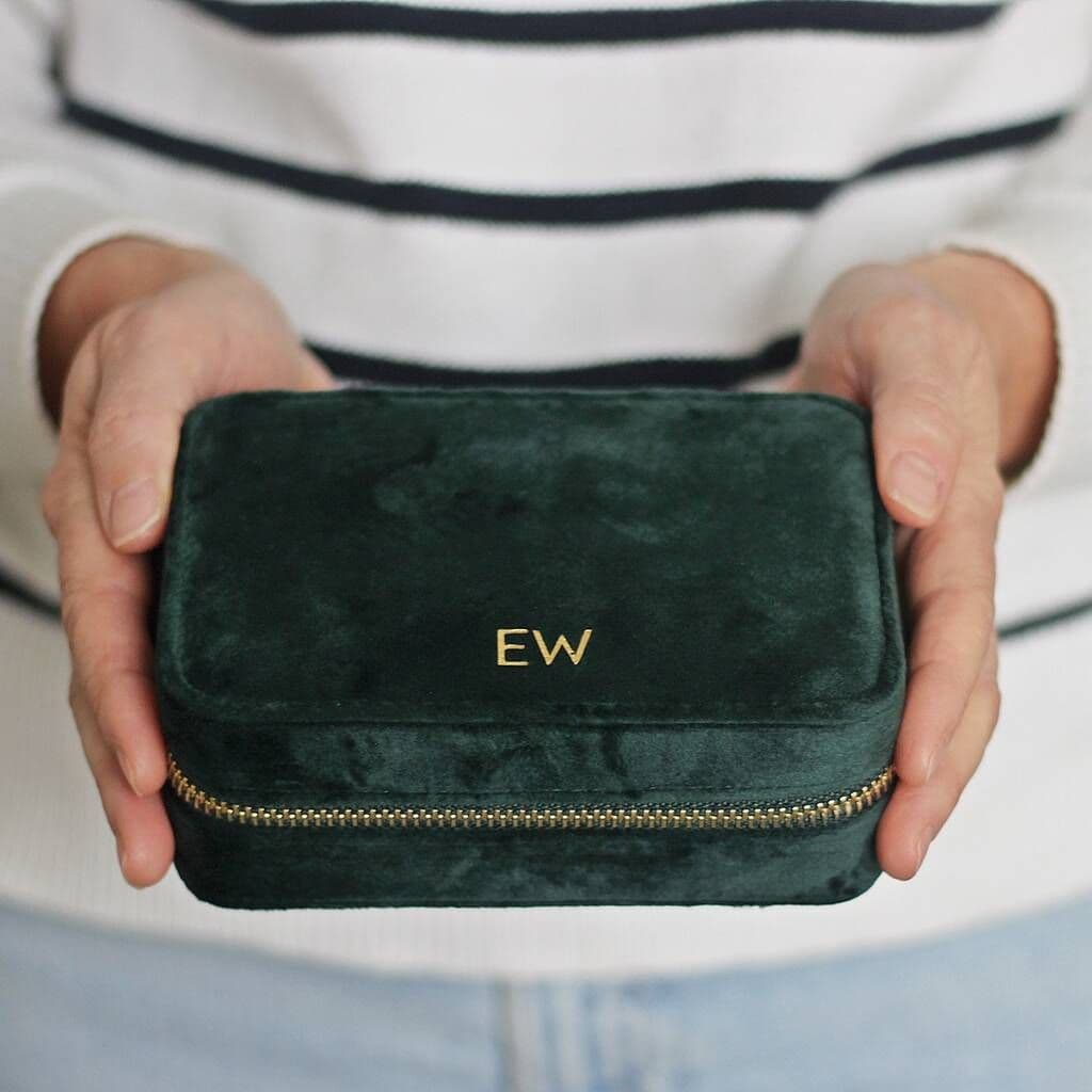 Green velvet pouch with gold zipper and initials held by a person wearing a striped shirt.