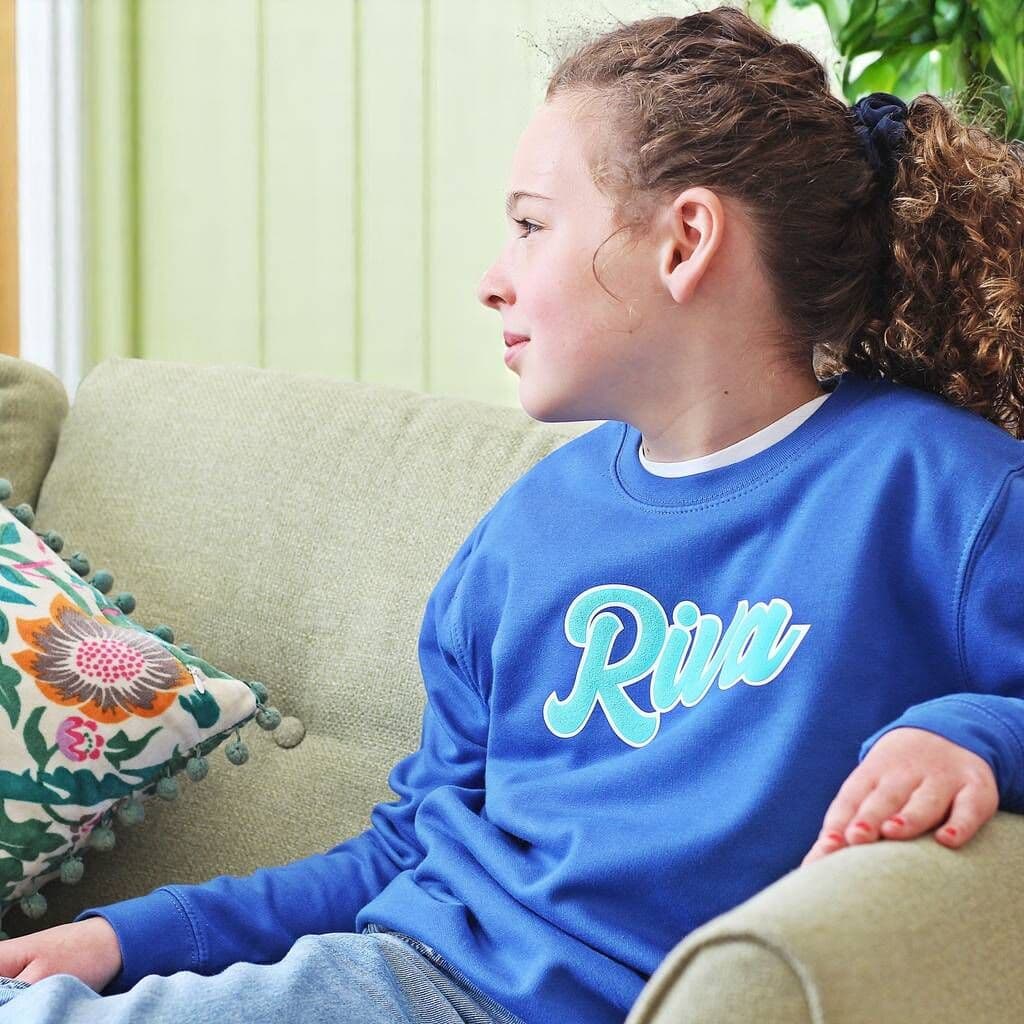 Child wearing a blue sweatshirt with 'Riv' on it, sitting on a couch.