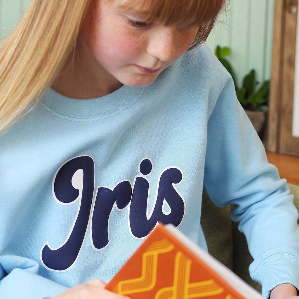 Child wearing a light blue sweatshirt with 'Iris' printed on it, holding a book.