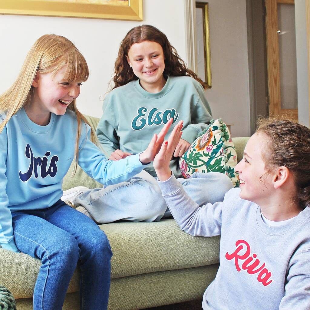 Three children sitting on a couch wearing sweatshirts with names printed on them.