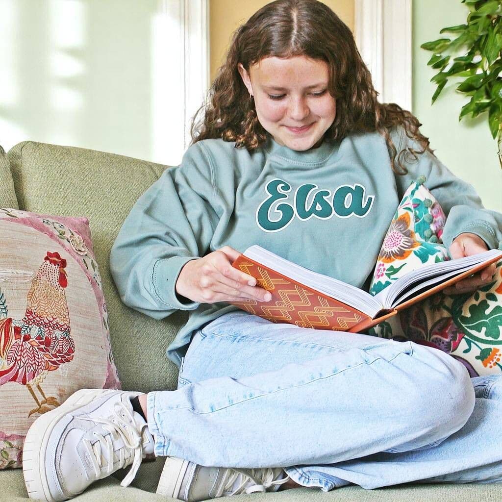 Person wearing a sweatshirt with 'Elsa' on it, sitting on a couch reading a book.