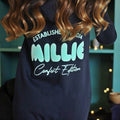 Navy onesie with 'MILLIE Comfort Edition' text on a blurred indoor background