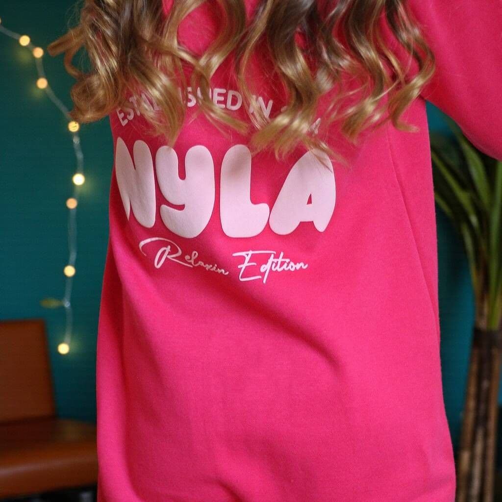 Person wearing a pink onesie with 'Nyla' printed on it against a teal wall with string lights.