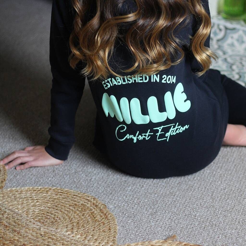 Person wearing a black Onesie with 'MILLIE Comfort Edition' text on the back, sitting on a carpeted floor.