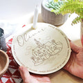 Personalized wooden plate with engraved design and text, held by a hand on a table with plants in the background.