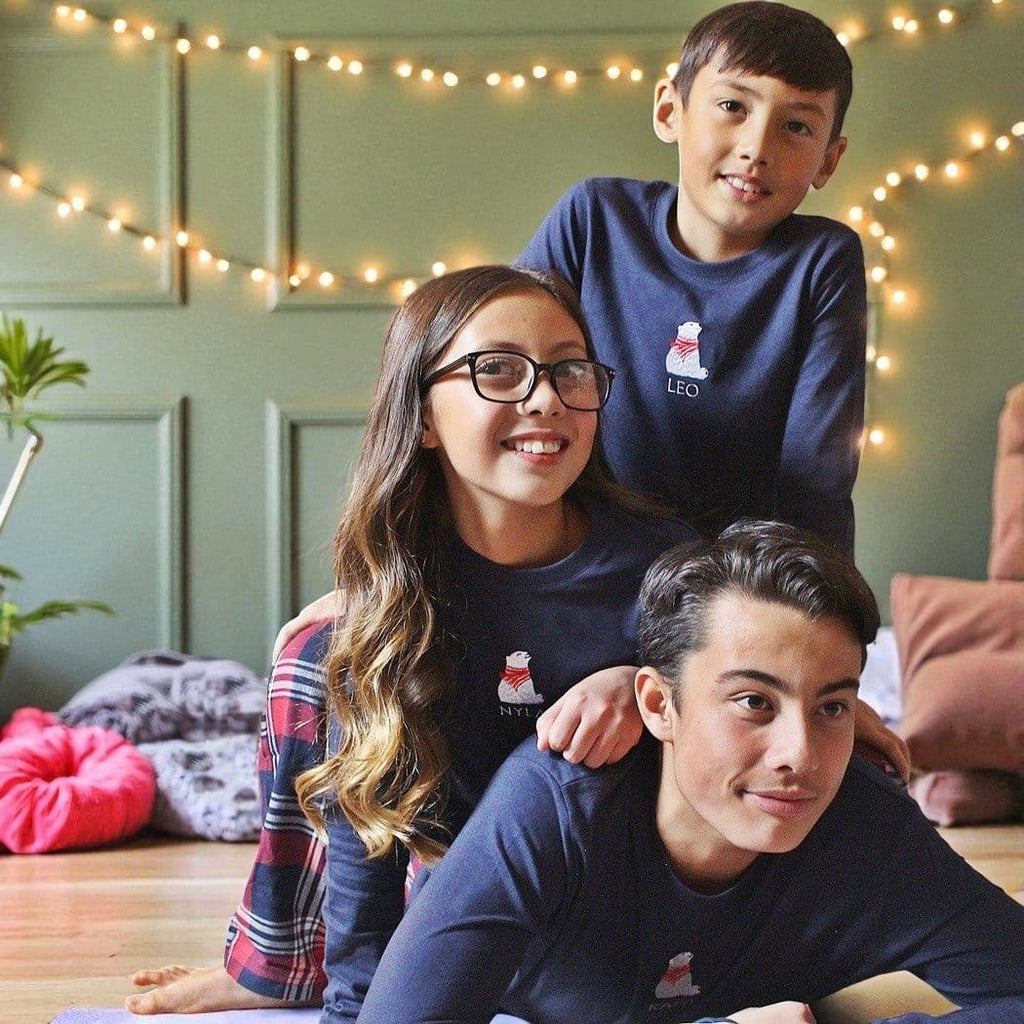 Three children wearing matching navy blue shirts with a logo, sitting on the floor with string lights in the background.