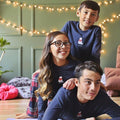 Three children wearing matching navy blue shirts with a logo, sitting on the floor with string lights in the background.