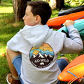 Child wearing a hoodie with 'Go Wild' design, sitting next to kayaks outdoors.