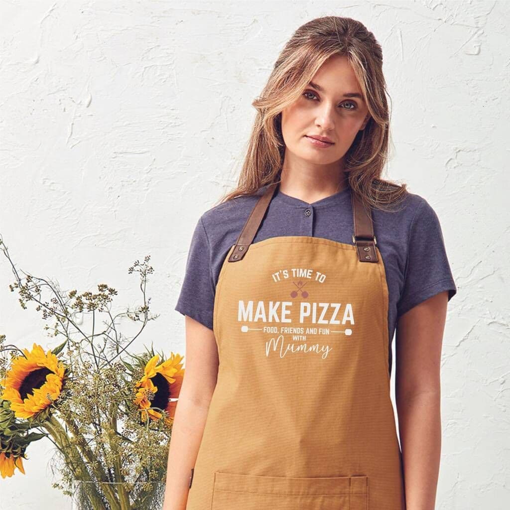 Woman wearing a mustard yellow apron with text, standing against a white wall with sunflowers.