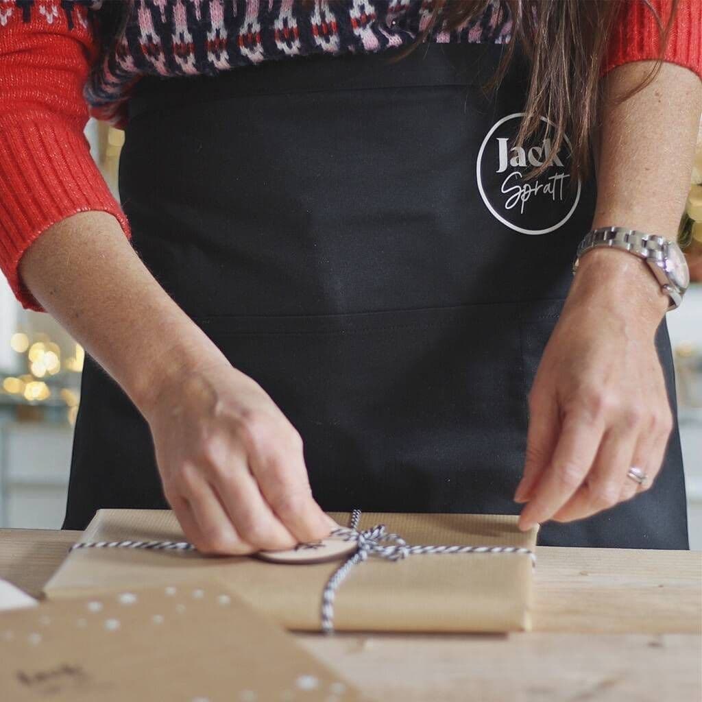 Person in a black apron with 'Jack Spratt' logo wrapping a gift.