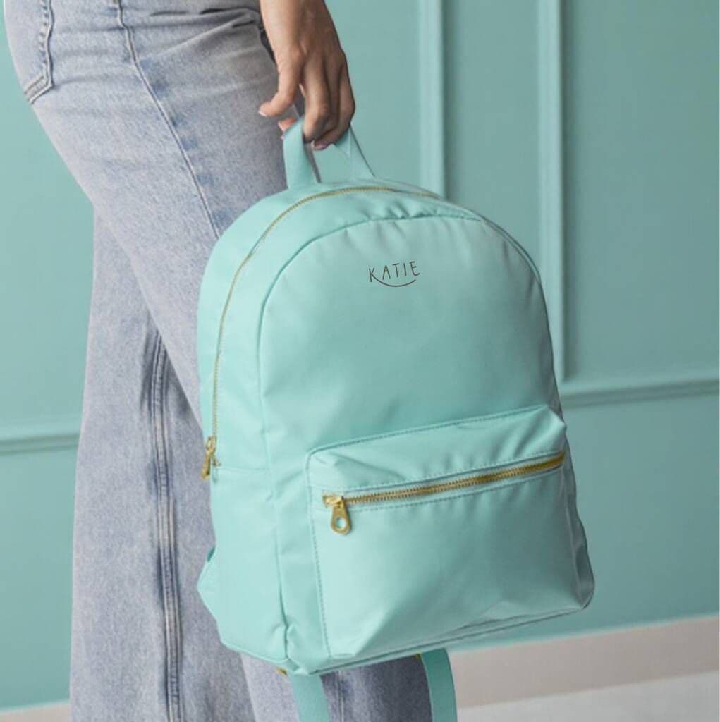 Mint green backpack with 'Katie' branding held by a person against a teal wall.