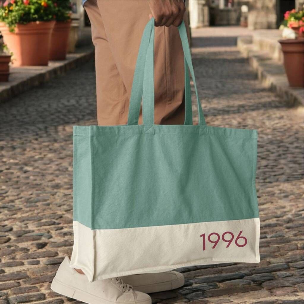 Person holding a green and white tote bag with '1996' on a cobblestone street.
