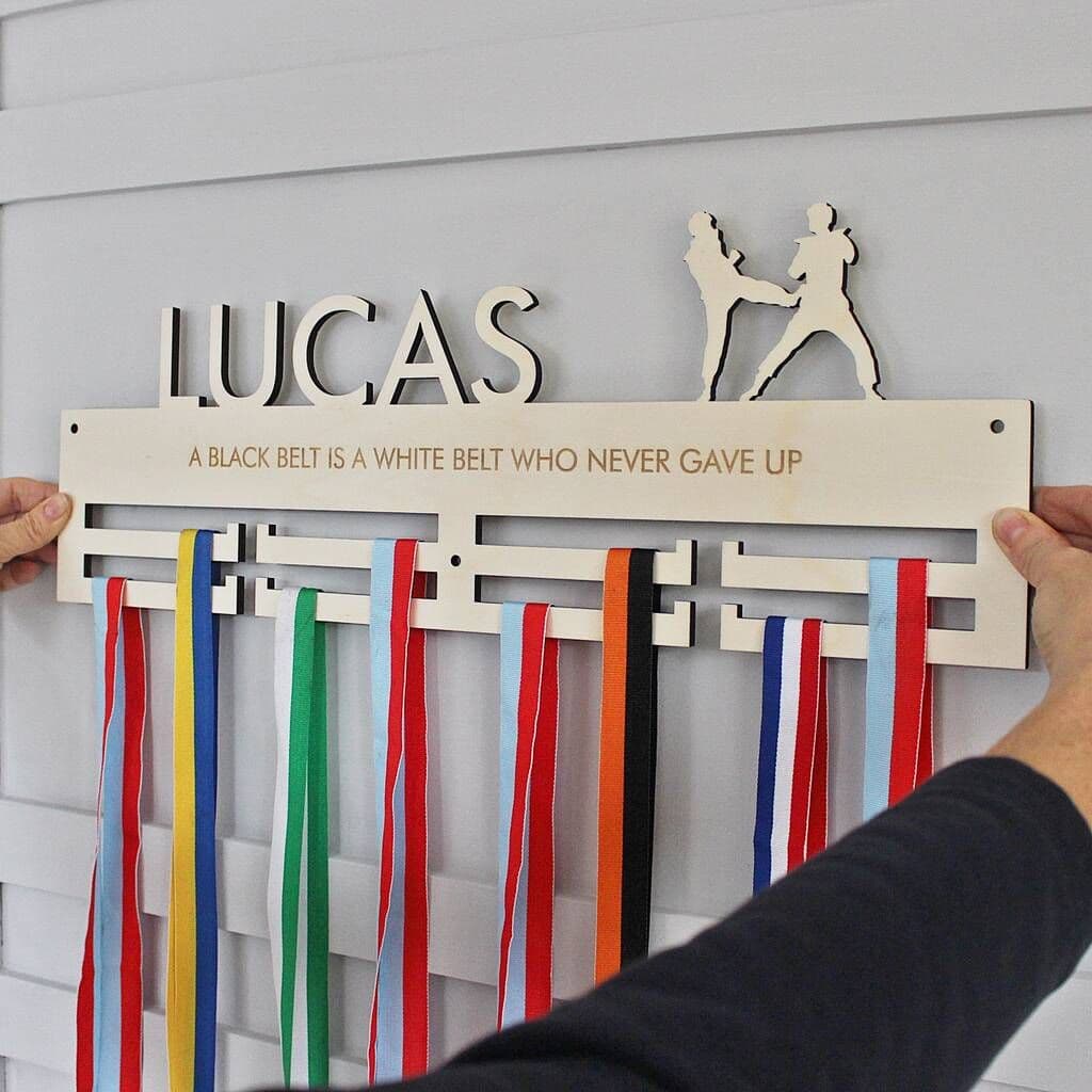 Wooden belt display with colorful ribbons and engraved text, held by two hands against a light gray background.