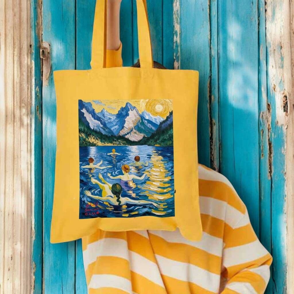 Yellow tote bag with a painting of a mountain lake design, held against a blue wooden background.