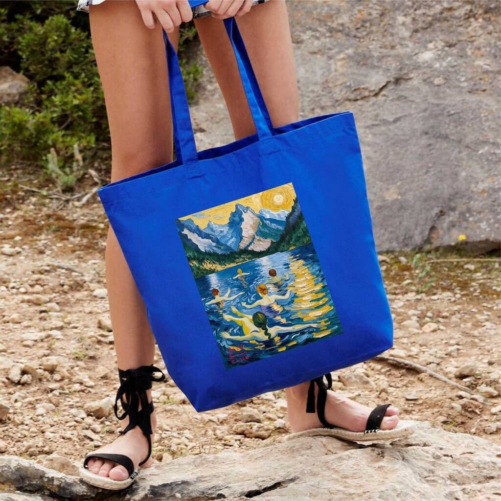Person holding a blue tote bag with a painting design outdoors.